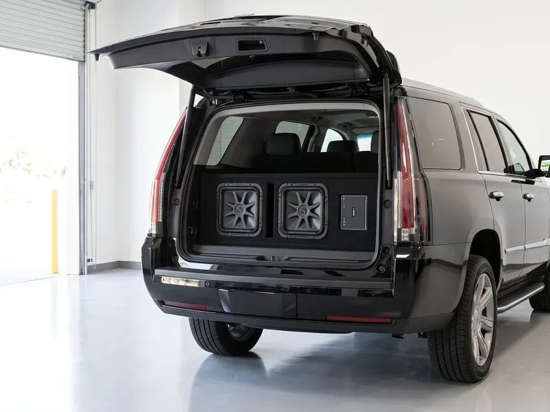 Black Cadillac Escalade with the tailgate open showing two Kicker subwoofers in the trunk